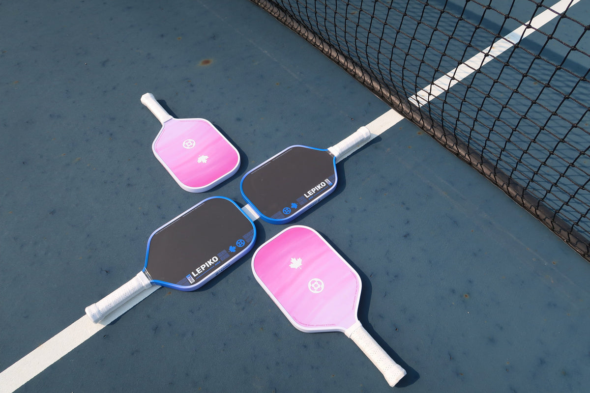 Four pickleball paddles arranged in a square on a blue pickleball court near the net. Two paddles are pink with white handles and blue trim, and two are dark gray/black with white handles and blue trim.